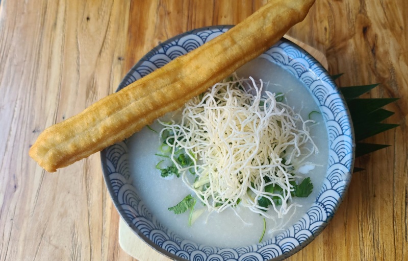 Jok khreung nai is a classic Thai breakfast of hearty rice porridge featuring ground pork and liver, topped with ginger, scallion, cilantro, fried rice noodles, and Chinese-style doughnut.