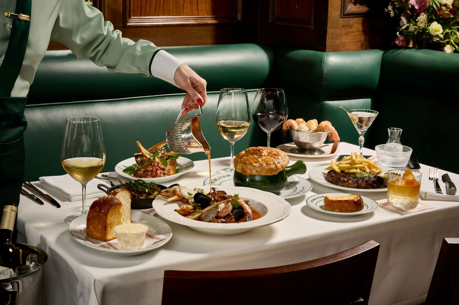 A table filled with gourmet dishes and drinks, including seafood, pie, bread, and steak. A hand pours sauce, conveying elegance and indulgence.