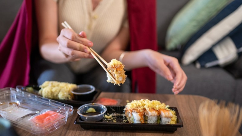 A woman holding chopsticks picks up a piece of sushi from a takeout container