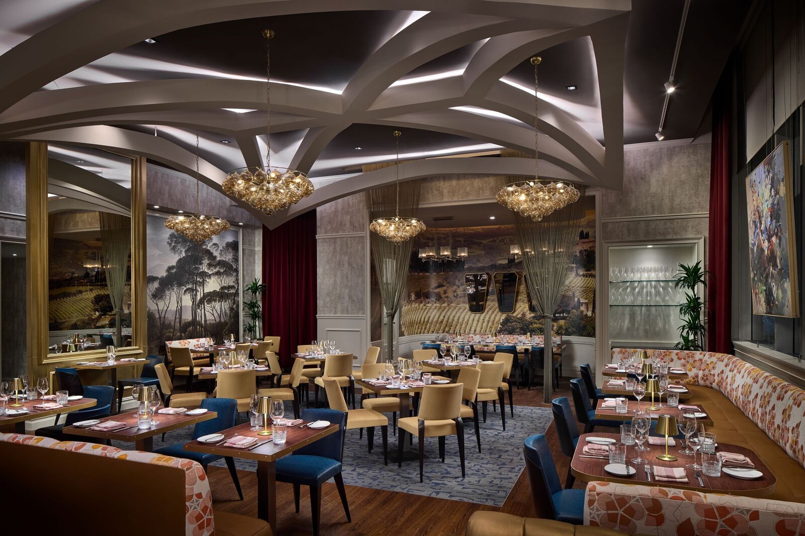 an empty high end restaurant with chandeliers, gold mirrors, and large image of trees and a field