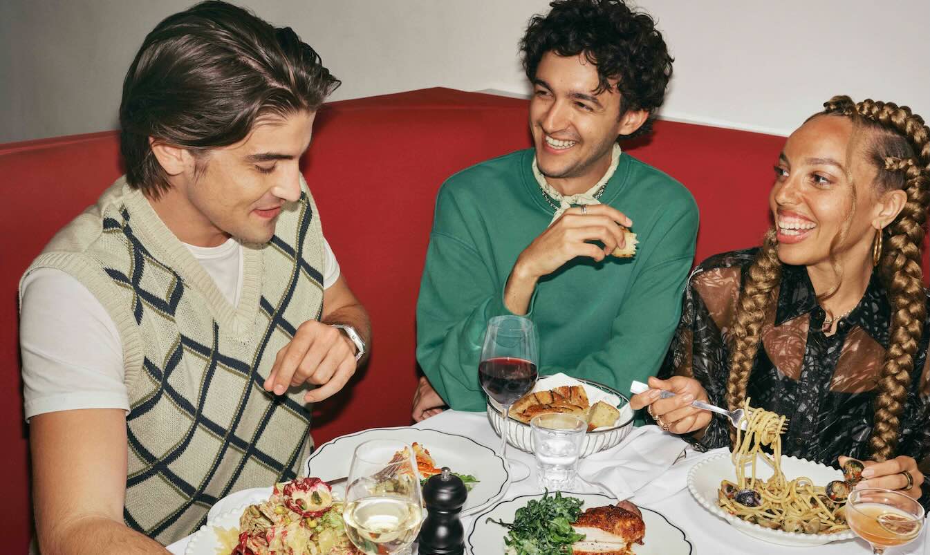 two men and a woman eating food with glasses of wine in red booth