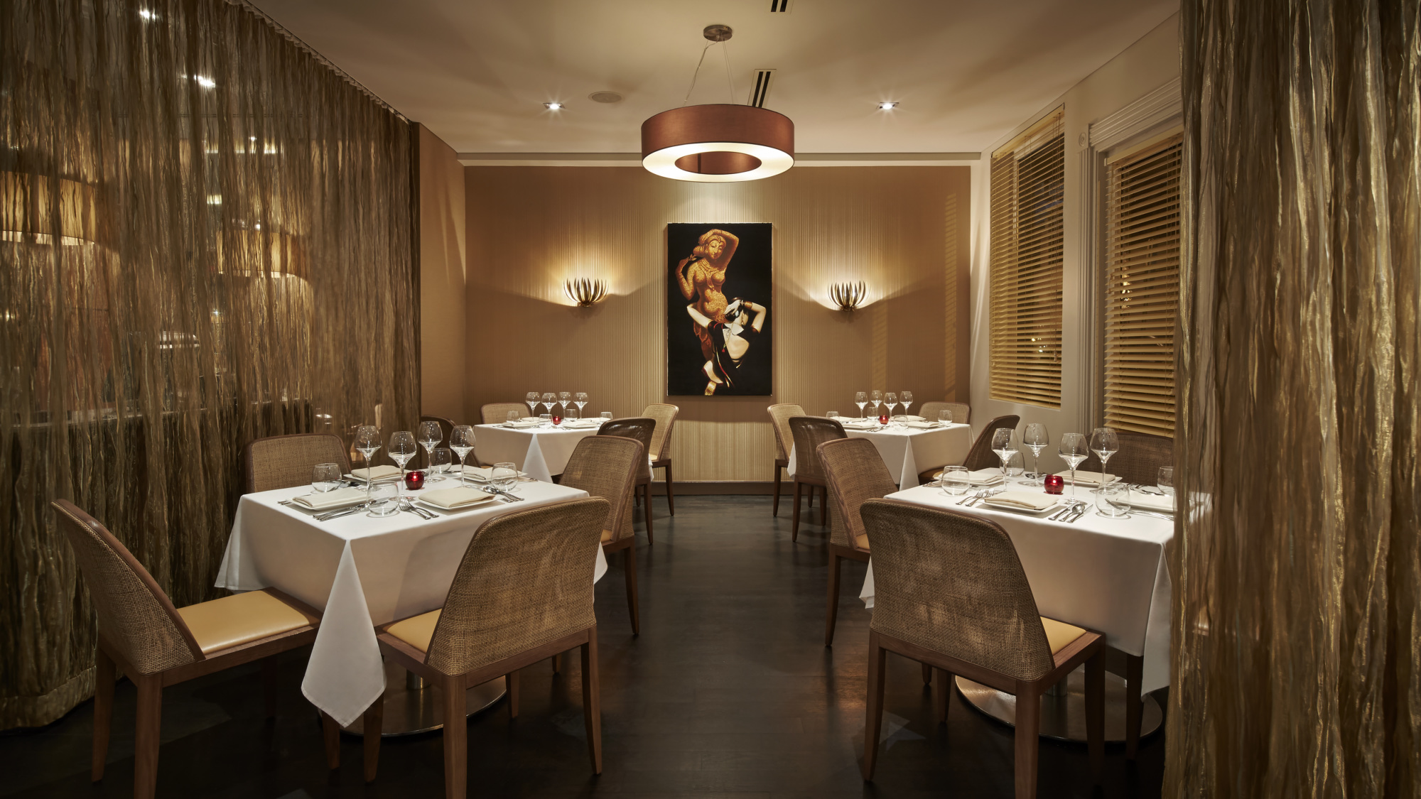 Elegant restaurant dining area with tables set for four, wicker chairs, white tablecloths, gold drapes, and a painting on the back wall under modern lighting.