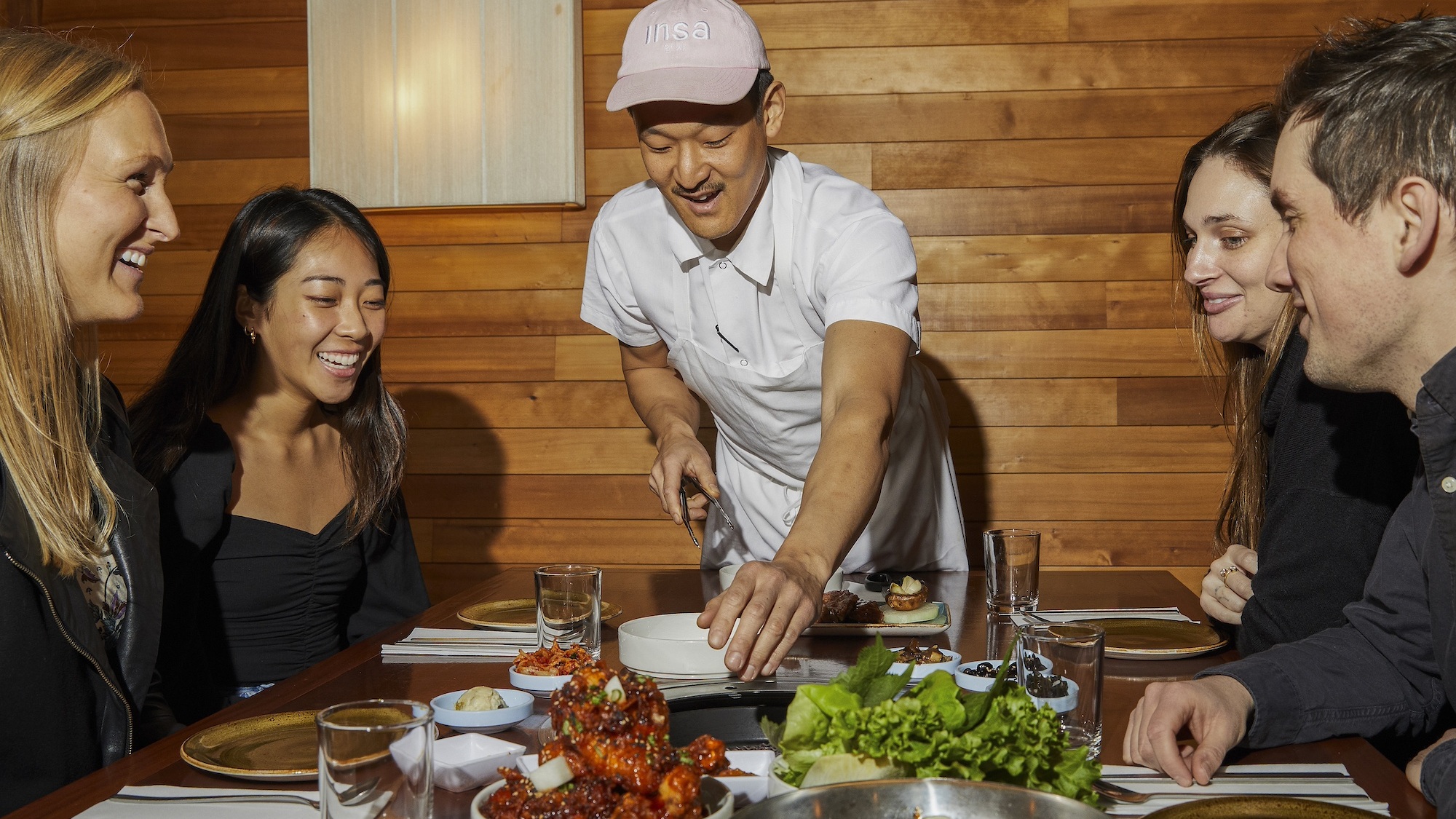 A group of people seated at a table, engaged in conversation with a chef presenting a dish.