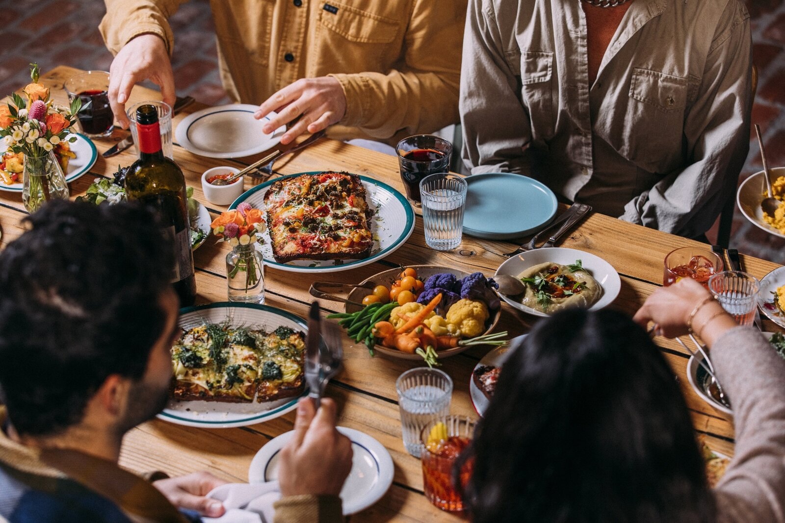 A restaurant table filled with dishes of food, plates, and silverware with seated diners' hands