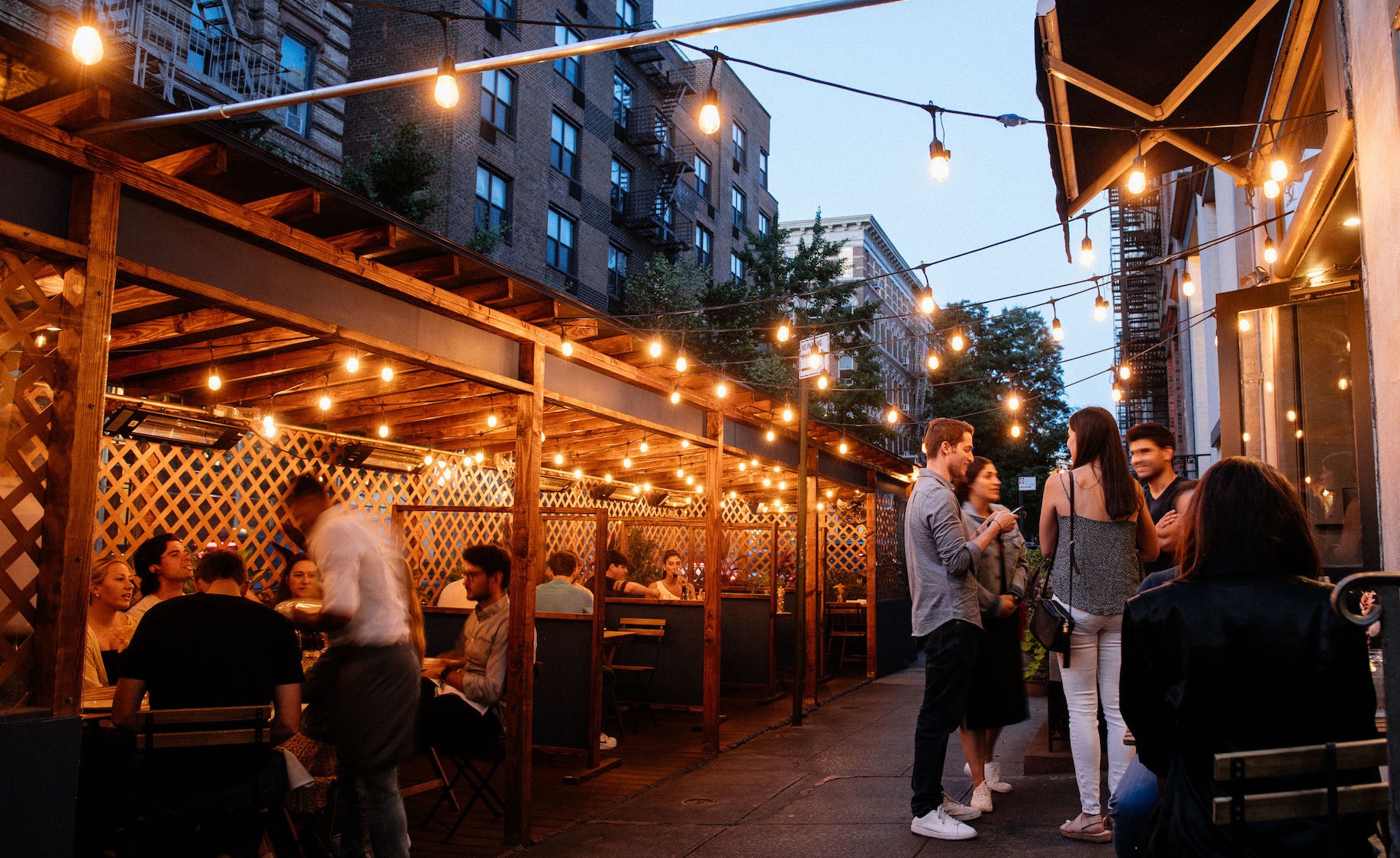 Diners stand outside at dusk under string lights near an outdoor patio