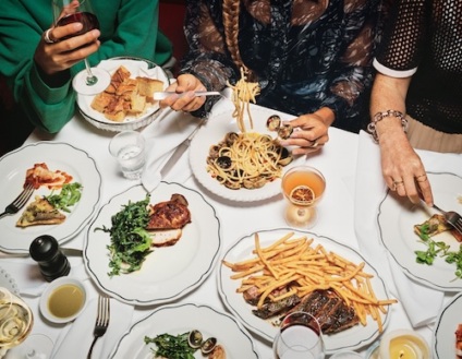 Overhead view of diners sharing pasta, steak, fries, and drinks at a lively restaurant table.